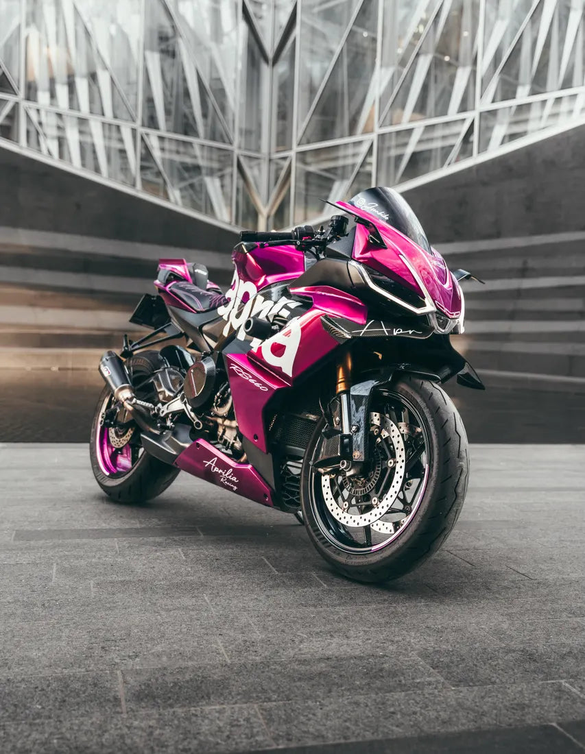 Aprilia RS660 rose avec stickers aprilia blancs, devant un bâtiment vitré